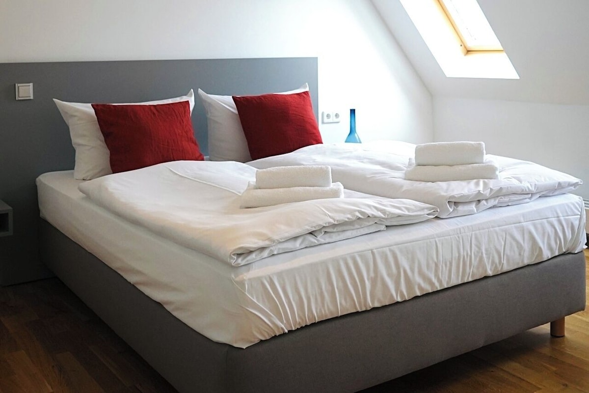 A modern bedroom with a sloped ceiling, a skylight, a bed with white linens, and a grey headboard.