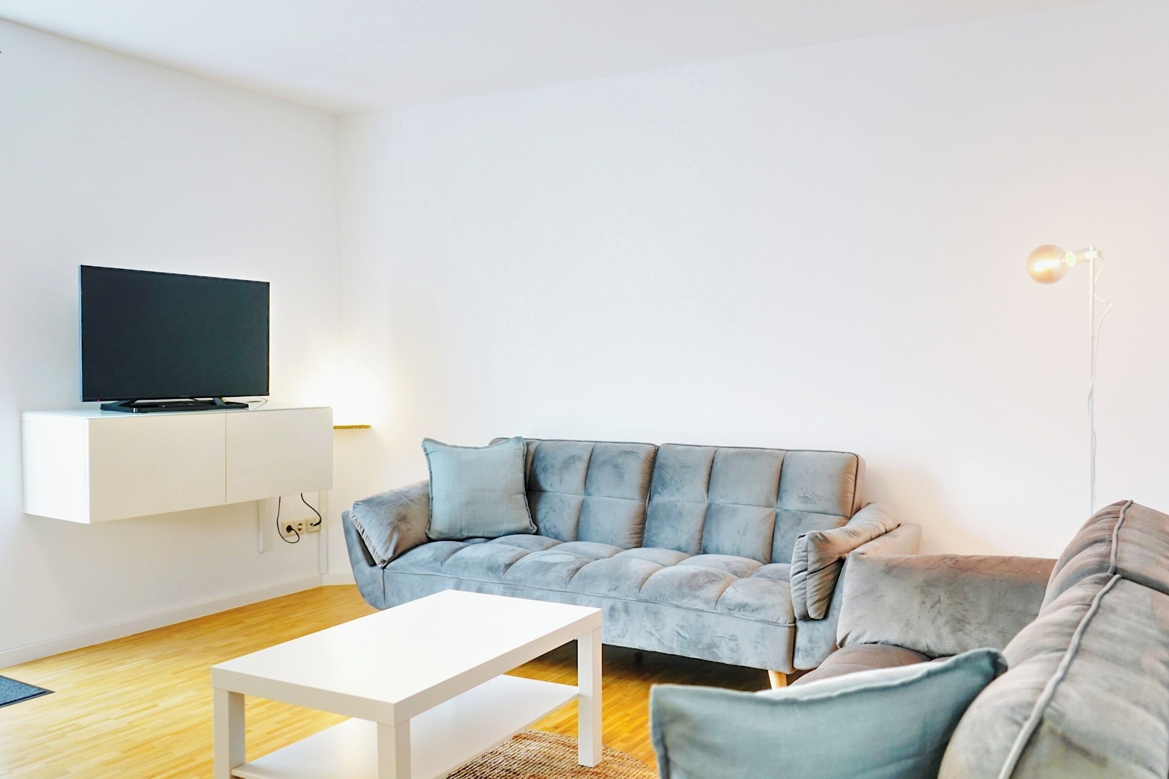 A modern living room with a flat-screen TV on a white cabinet, a grey sofa, a white coffee table, and a floor lamp.