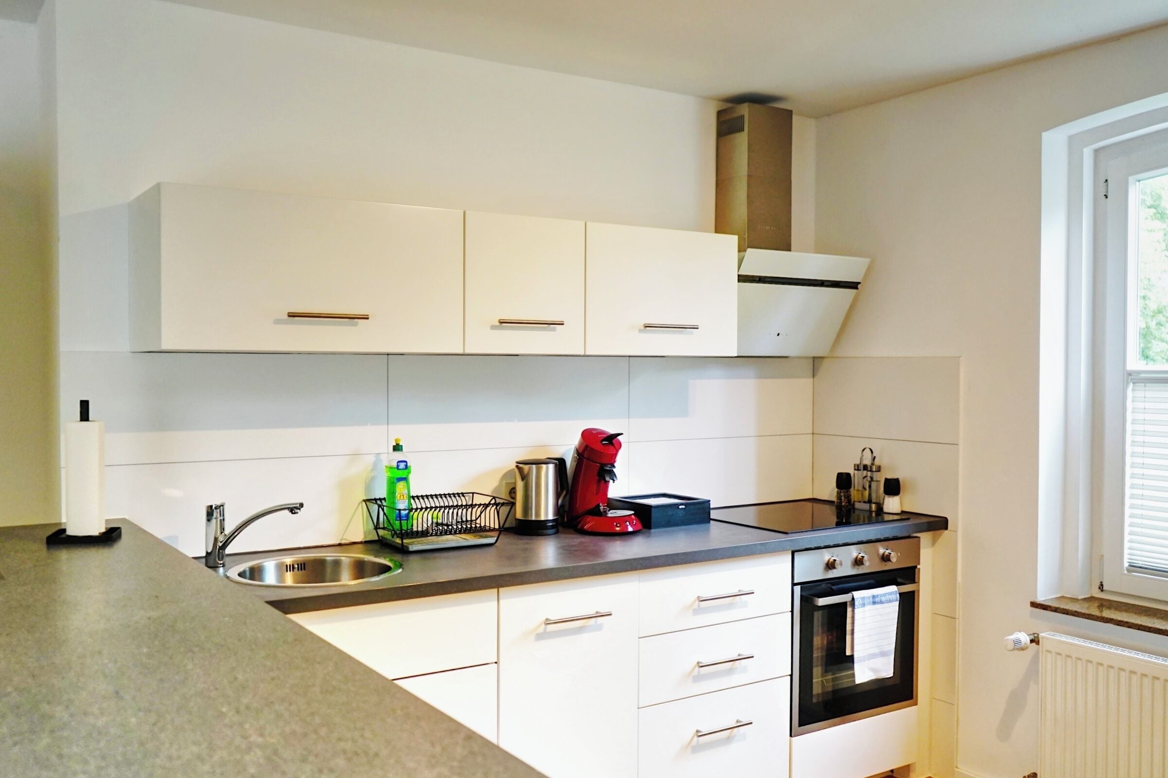 A modern kitchen with white cabinets, a stainless steel sink, a red kettle, and a built-in oven.