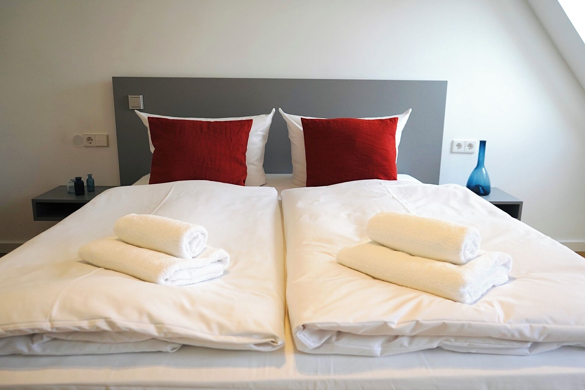 A hotel room with two beds, each with white linens and rolled towels, a grey headboard, and a small bedside table with a blue vase.