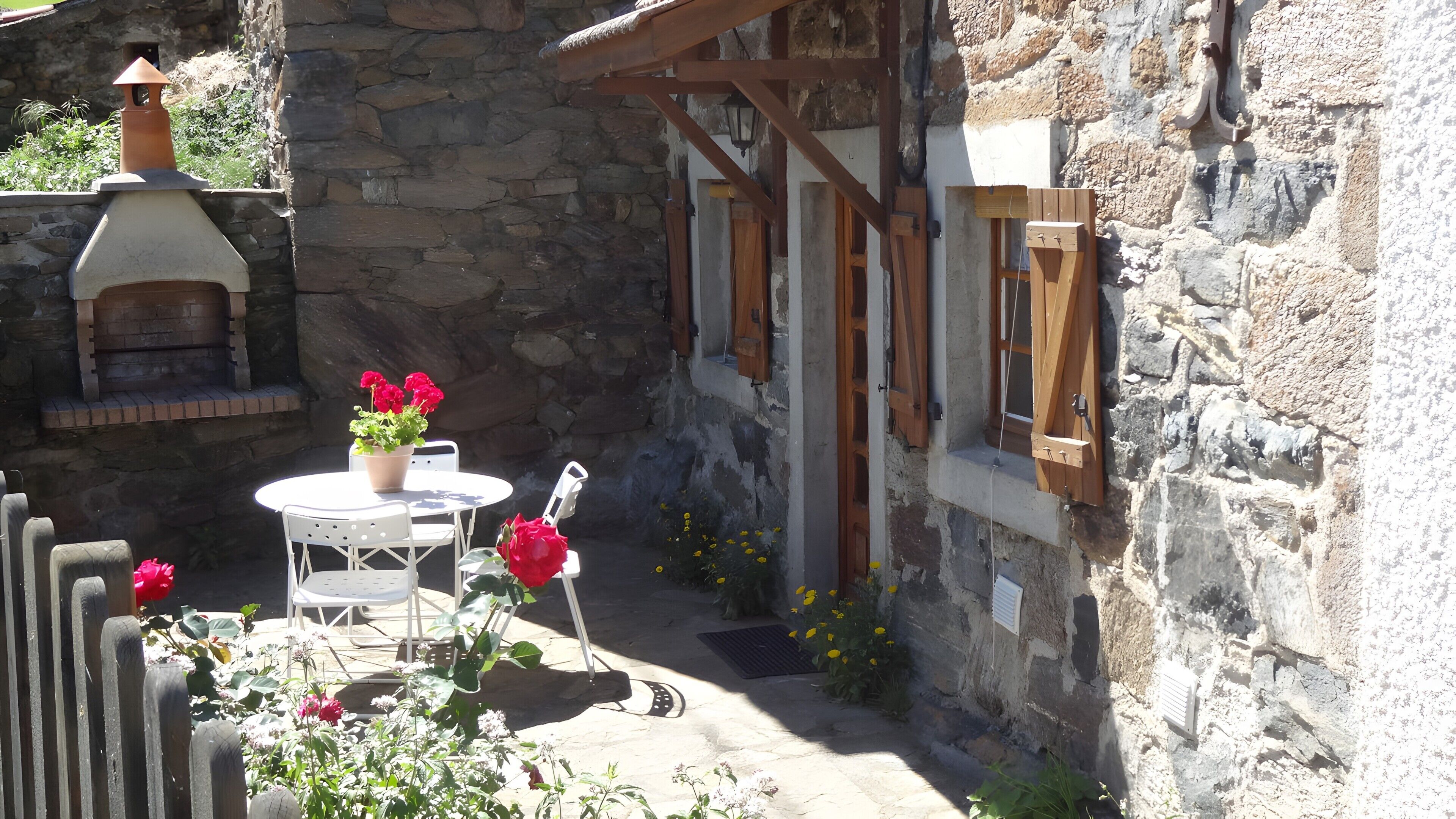 Un bâtiment en pierre doté d’un petit patio comprenant une table ronde et des chaises, un pot de fleurs avec des fleurs rouges et une structure en bois avec des fenêtres.