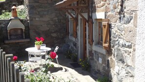 A stone building with a small patio area featuring a round table and chairs, a flower pot with red flowers, and a wooden structure with windows.