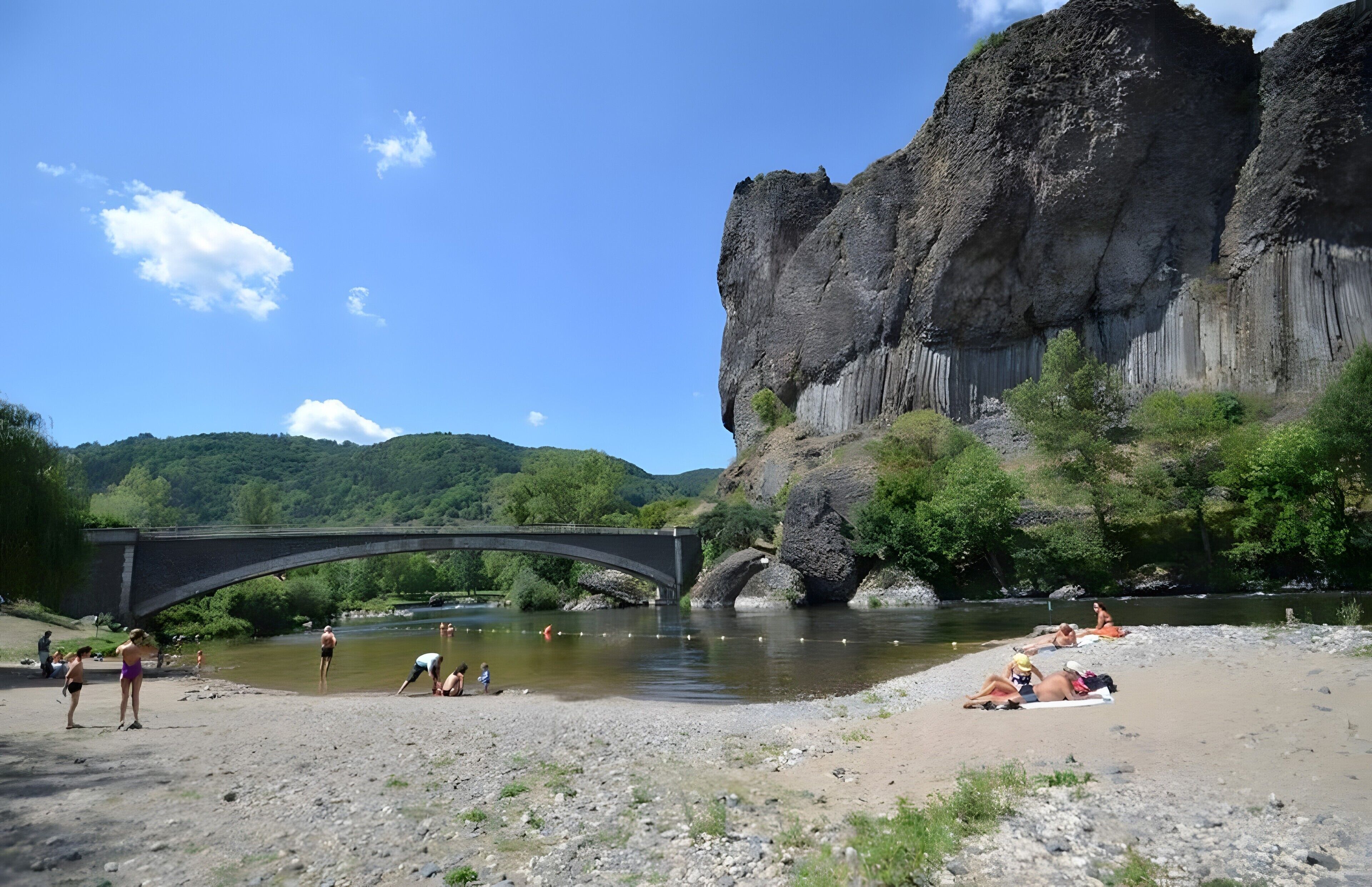 Une rivière traversée par un pont, des personnes se détendant sur la rive et une grande formation rocheuse.