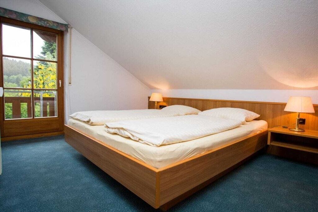 Cama de solteiro com cabeceira de madeira em quarto com teto inclinado, janela com vista para área verde e duas luminárias de cabeceira.