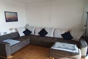 A living room with a sectional sofa, a small table, and a wall-mounted lamp. - Holiday apartment with 1 bedroom (Wilhelmshaven)