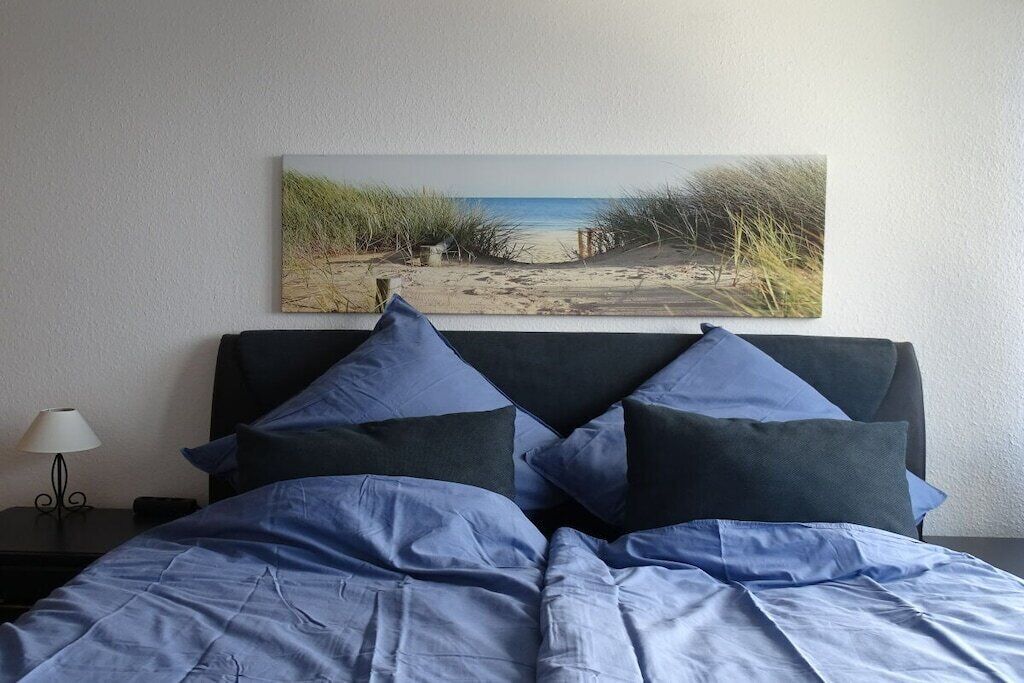 En seng med lyseblåt dynebetræk og puder, et natbord med lampe, og et maleri med et strandmotiv på væggen.