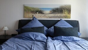 Ein Bett mit blauer Bettdecke und Kissen, ein Nachttisch mit Lampe sowie ein Strandbild an der Wand.