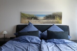 En säng med ett blått överkast och kuddar, ett sängbord med en lampa och en tavla med strandmotiv på väggen.