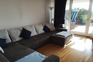 Un salon moderne avec un canapé d’angle, un petit pouf, une lampe sur pied et une vue sur l’extérieur grâce à une porte-fenêtre coulissante.
