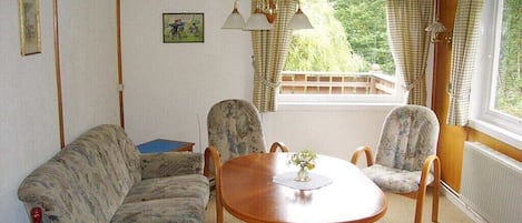 Una habitación con una mesa de comedor de madera, dos sillas estampadas, un sofá, un ventanal con cortinas y una botella sobre una mesa.