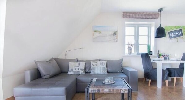 A modern living room with a grey sectional sofa, a coffee table, and a dining area with a white table and chairs. - Ordinger Dün Holiday Apartment 6 (St. Peter-Ording)