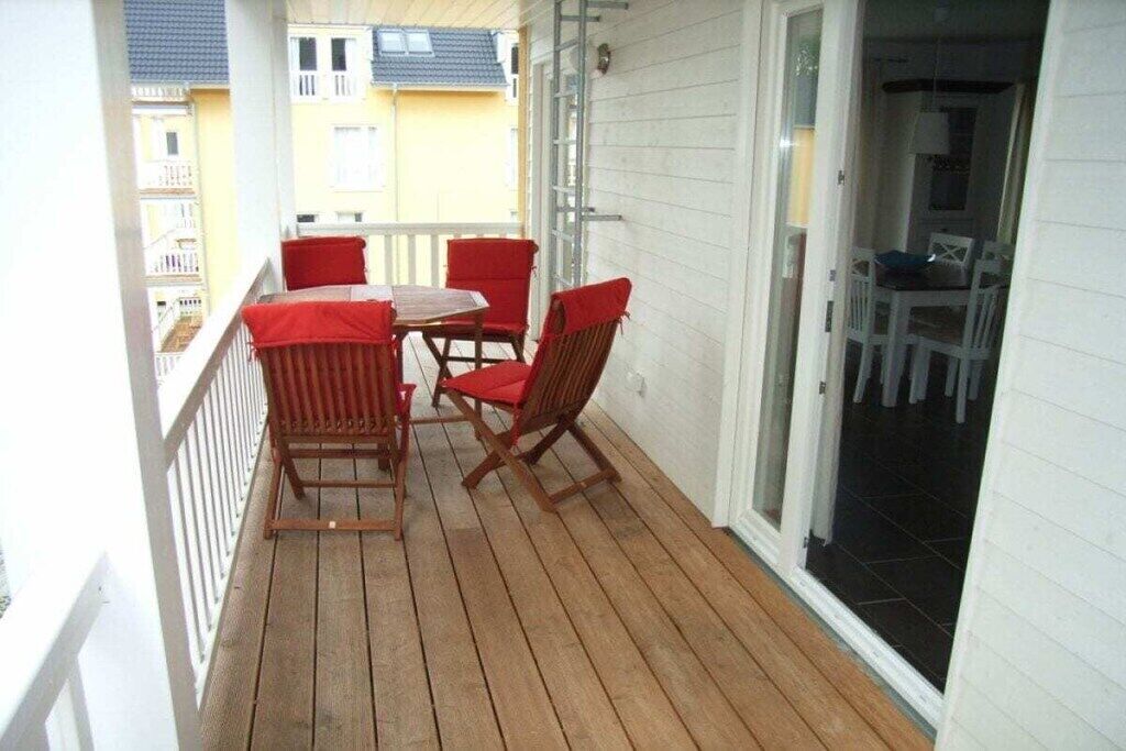 En træterrasse med et bord og stole, et hvidt gelænder og en glasdør, der fører ind i et rum.