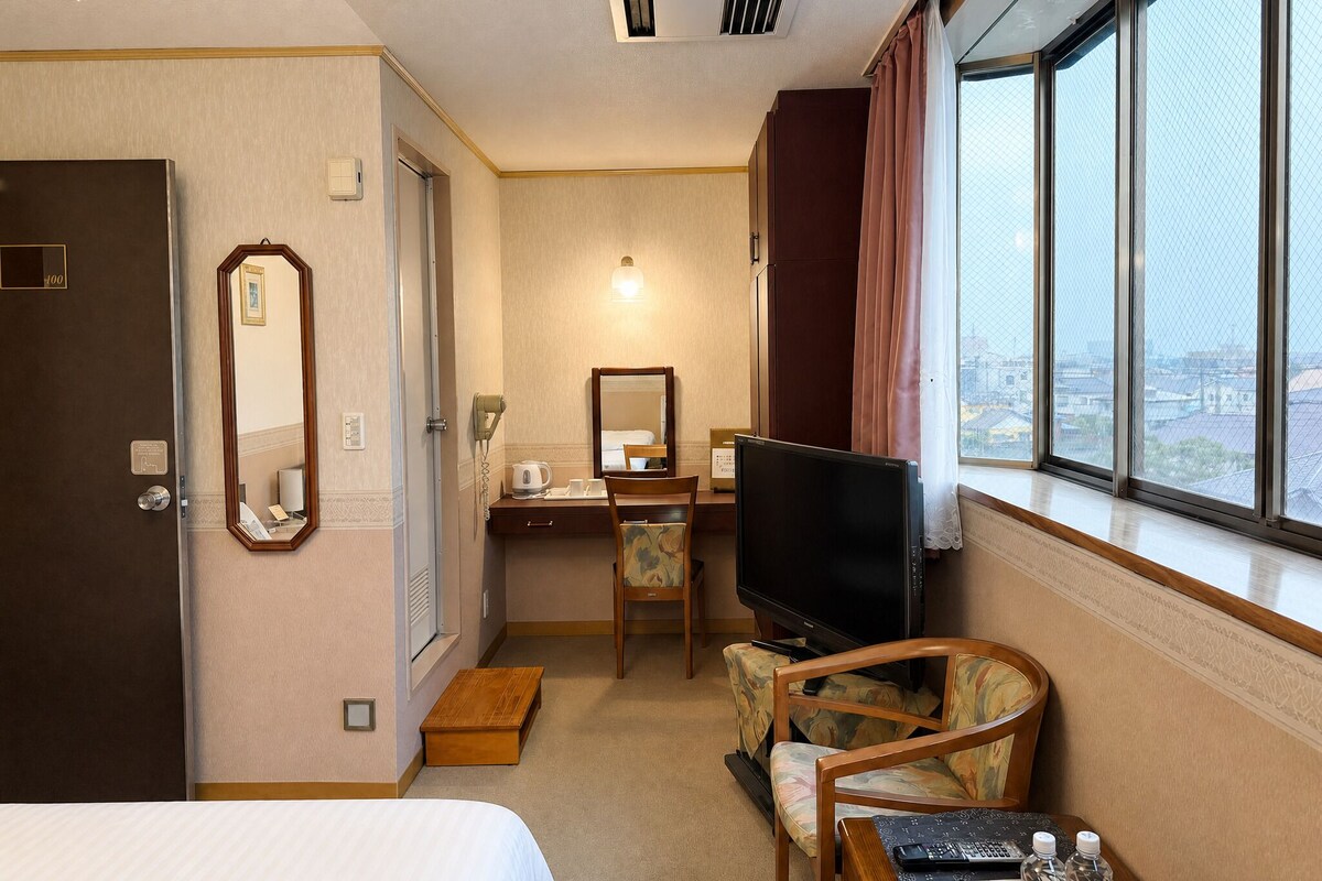 A hotel room with a bed, a desk with a chair, a television, and a large window with curtains.