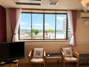 A hotel room with a large window, two chairs with cushions, a small table, a flat-screen TV, and a clock on the wall. - WASHUKAN HOTEL  (Gyoda)