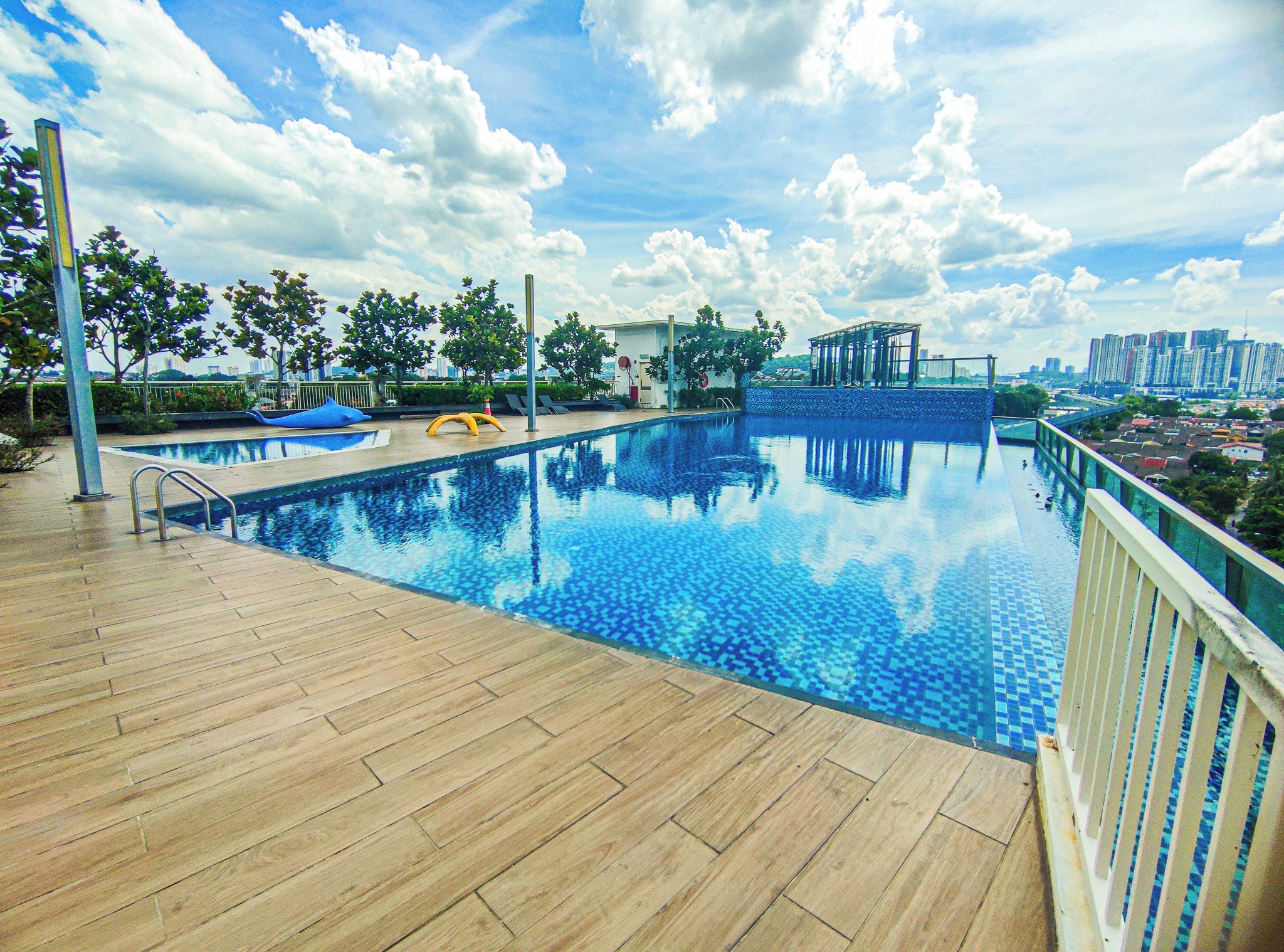 A rooftop infinity pool with a wooden deck, surrounded by trees and a cityscape view.