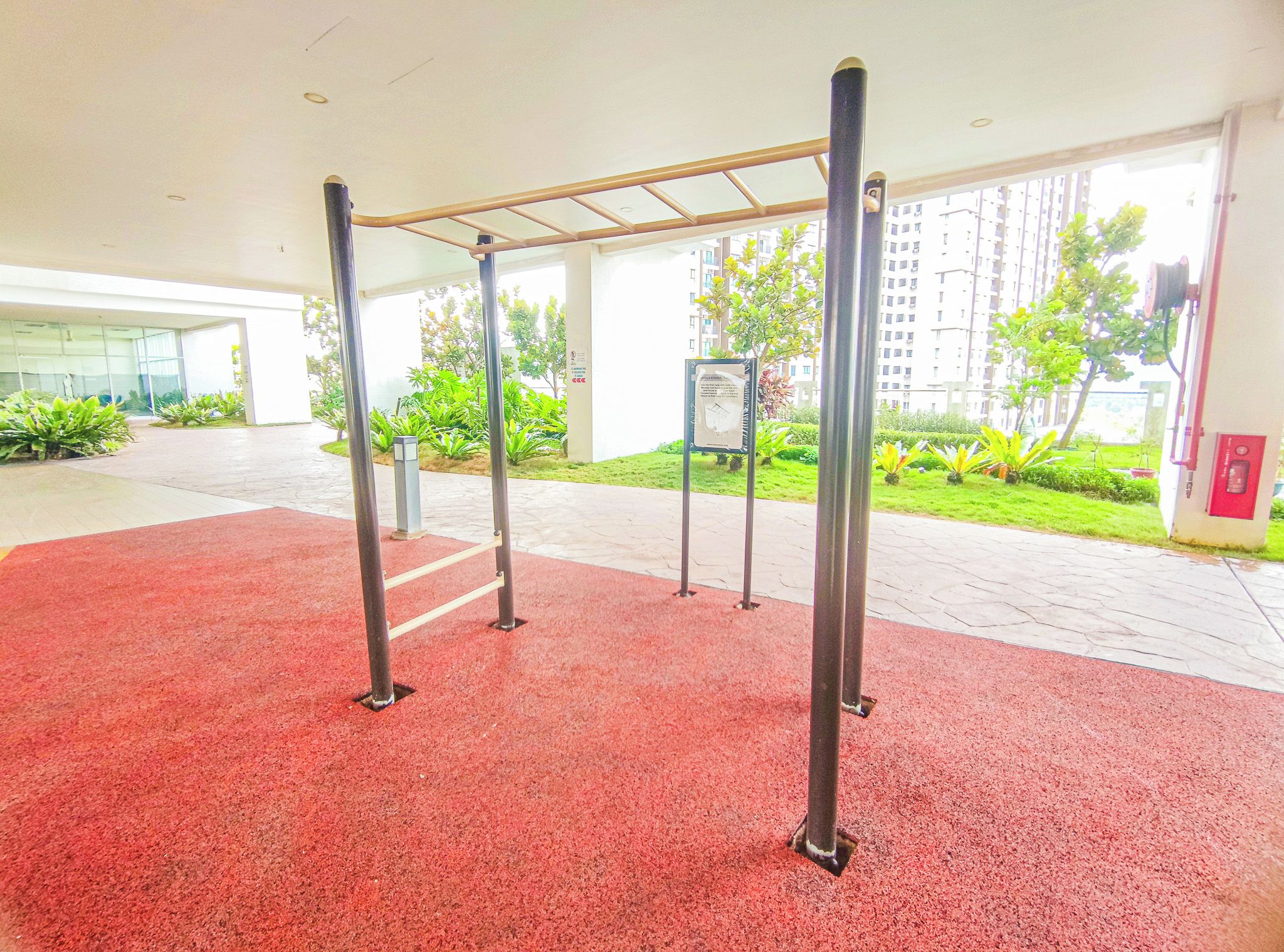An indoor gym with a pull-up bar structure, red flooring, and a view of a residential area with greenery and buildings.
