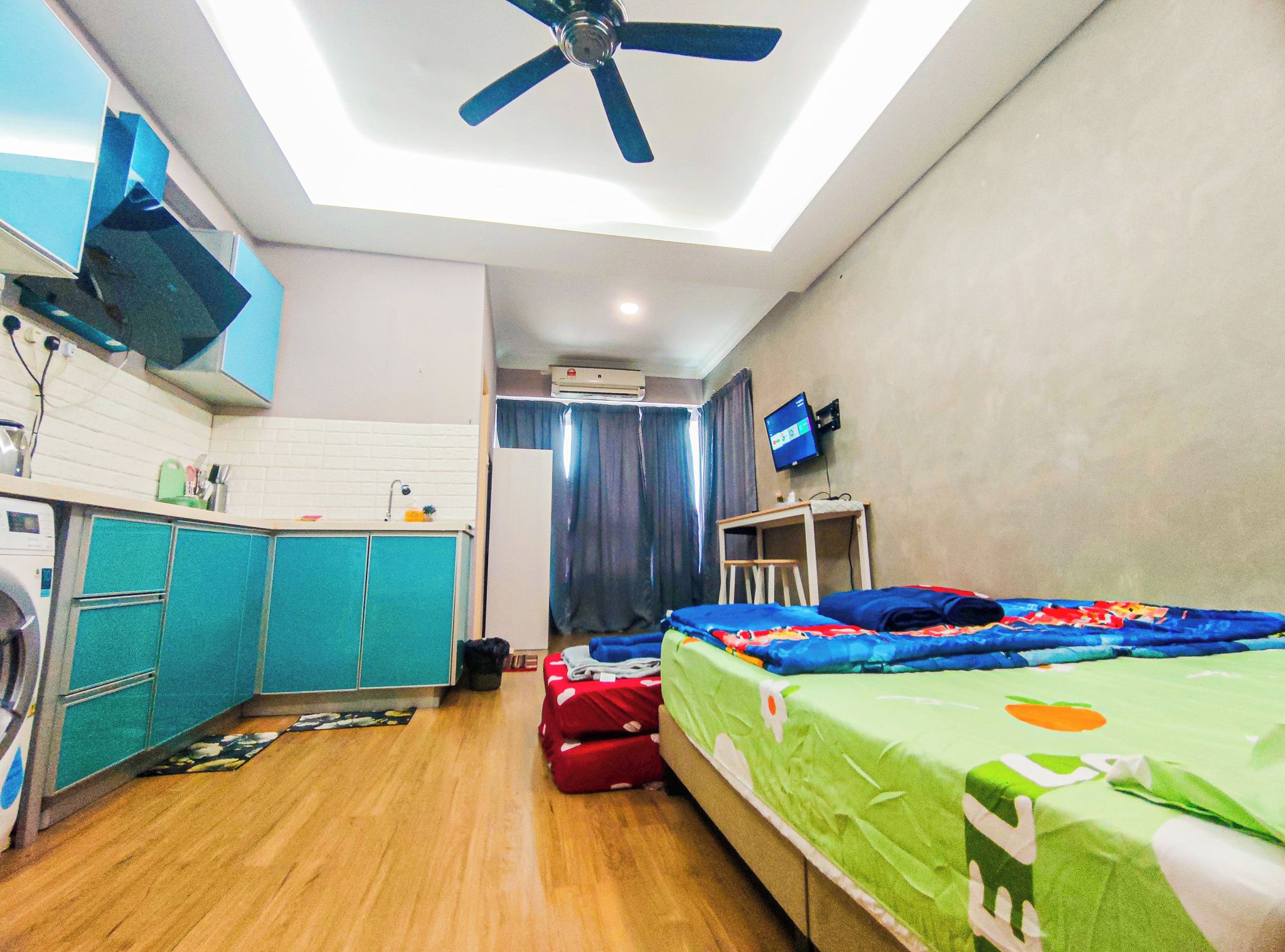 A compact hotel room with a bed, a desk, a ceiling fan, a TV, and a kitchenette with a washing machine.
