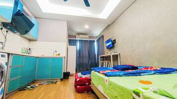 A compact hotel room with a bed, a desk, a ceiling fan, a TV, and a kitchenette with a washing machine.