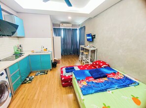 Una habitación compacta con kitchenette, una cama con colcha colorida, un escritorio con computadora y una lavadora.