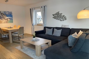 Ein modernes Wohnzimmer mit einem dunkelgrauen Sofa, einem Holztisch und einem Essbereich mit Tisch und Stühlen.
