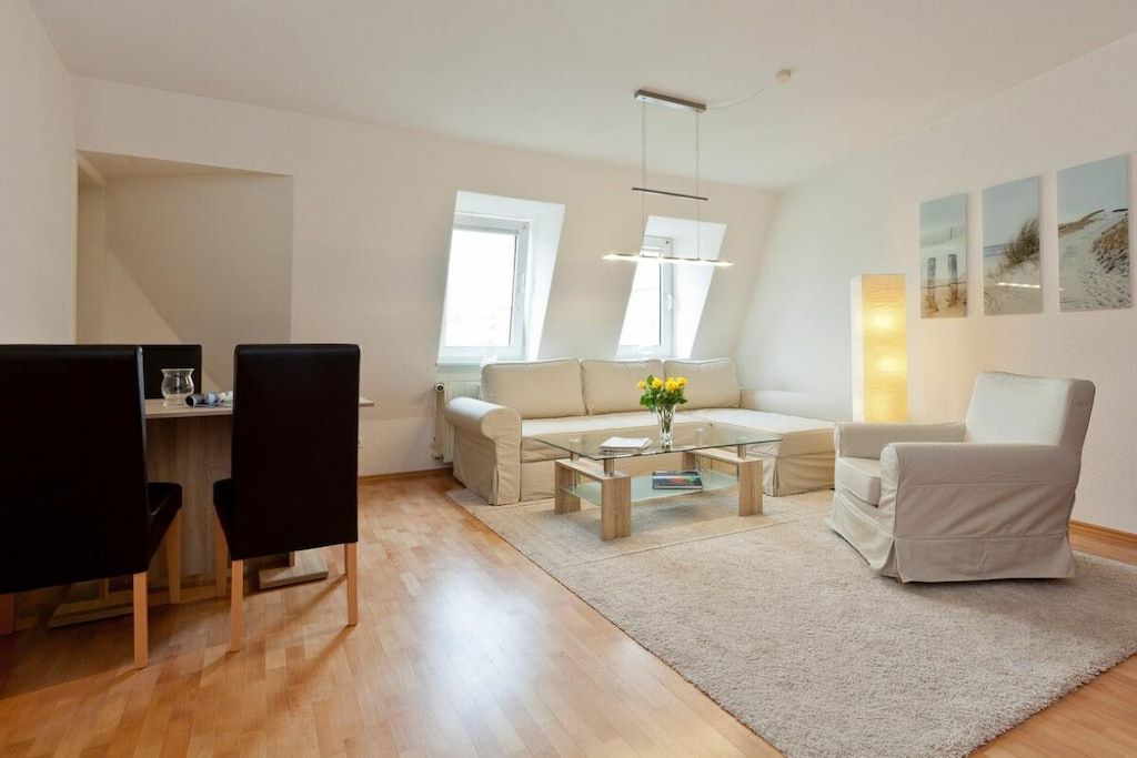 Una sala de estar moderna con sofá, mesa de centro y sillas. Hay un comedor con mesa y sillas. La habitación tiene ventanas amplias y una lámpara de techo.