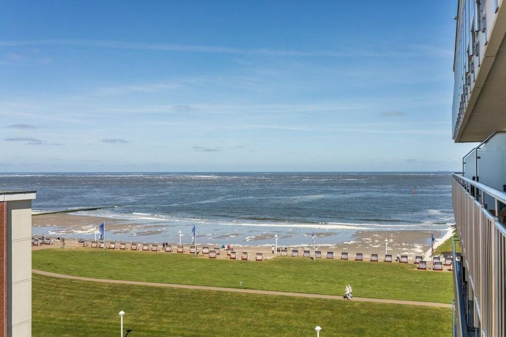 Vista de una playa con una fila de sillas y sombrillas, una zona verde y un edificio al frente.