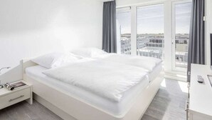 Et hotelværelse med en stor seng, et natbord med lampe, et skrivebord med stol og udsigt til byen gennem store vinduer.