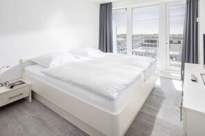 Habitación de hotel con una cama grande, una mesita de noche con lámpara, un escritorio con silla y vistas a la ciudad a través de amplios ventanales.