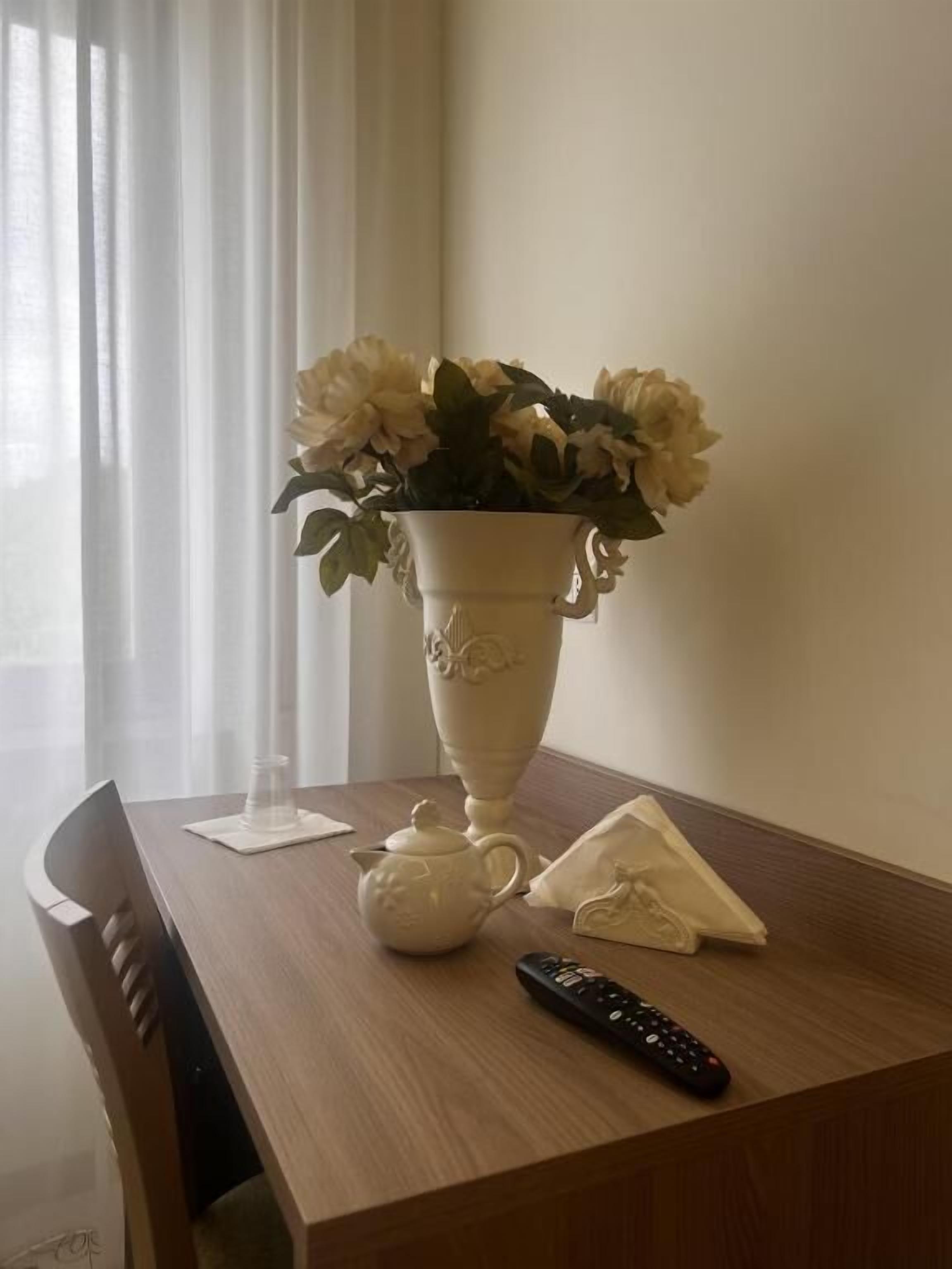 Una mesa con un jarrón blanco lleno de flores, un hervidor, un control remoto y una servilleta.