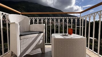 Een balkon met rieten meubilair, een ronde tafel en uitzicht op een bergachtig landschap.