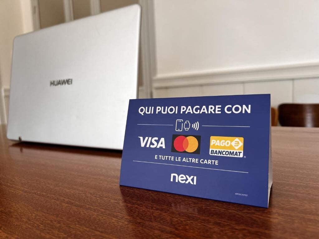 A blue payment card with Visa, MasterCard, and Bancomat logos, and the Nexi logo, placed on a wooden surface with a white laptop in the background.