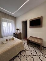 Una habitación de hotel con una cama, un banco, un televisor montado en la pared y una puerta que da a otra habitación.