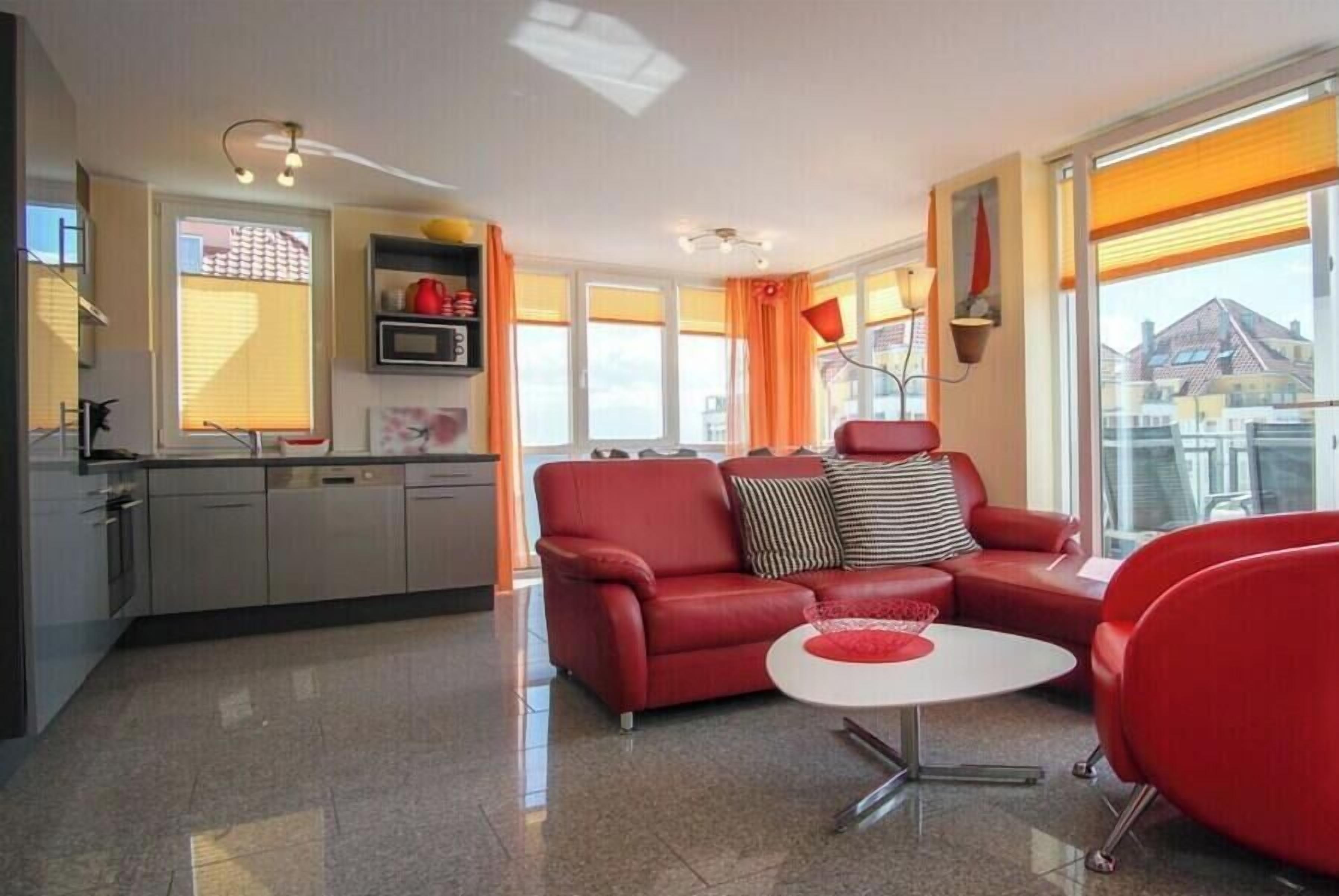Ein modernes Wohnzimmer mit einem roten Sofa, einem weißen Couchtisch und einer Küchenzeile mit grauen Schränken und einer Mikrowelle.