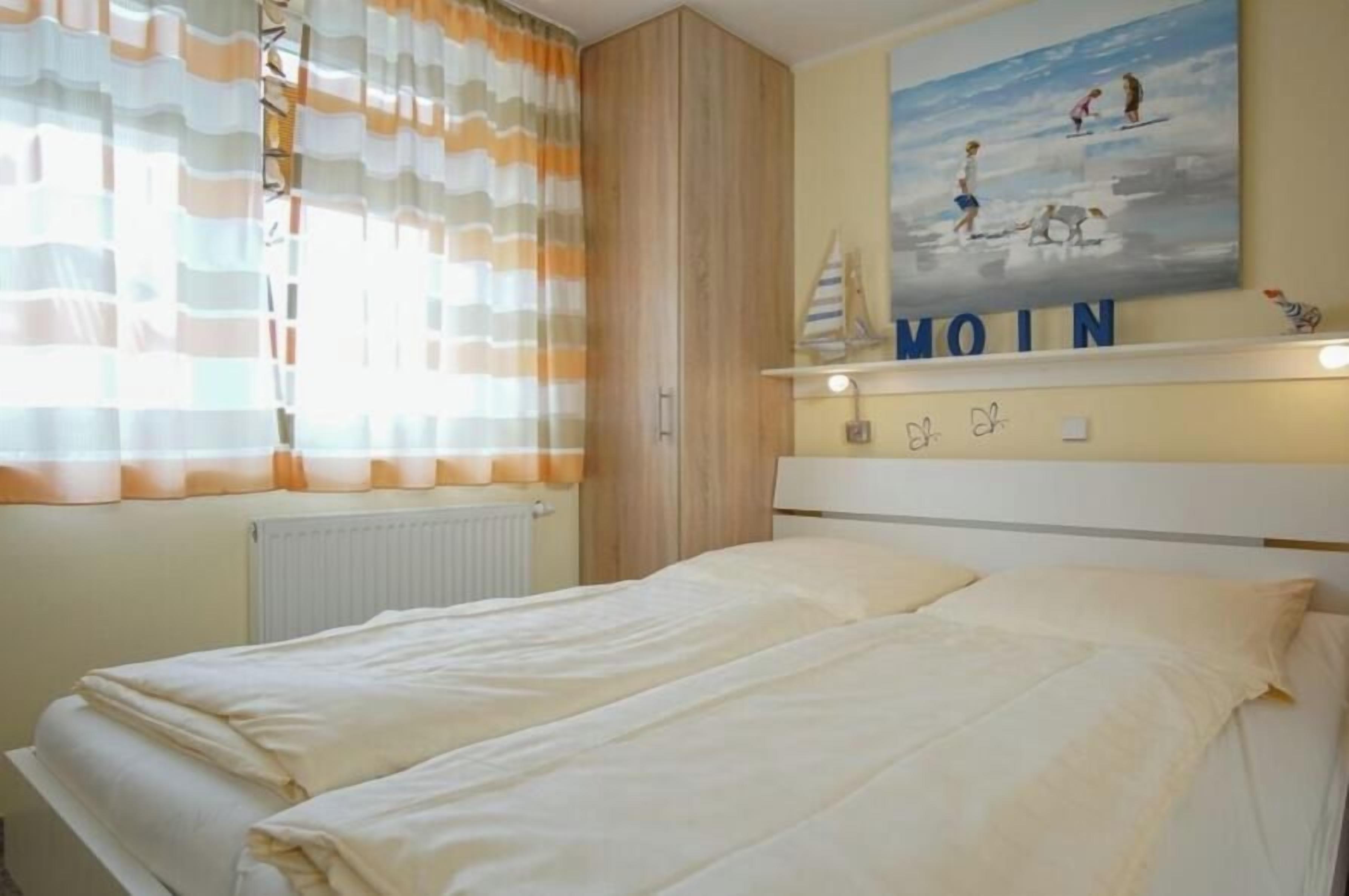 Quarto com cama, cortinas, radiador, prateleira com um modelo de veleiro e um quadro com a palavra 'MOIN' na parede.