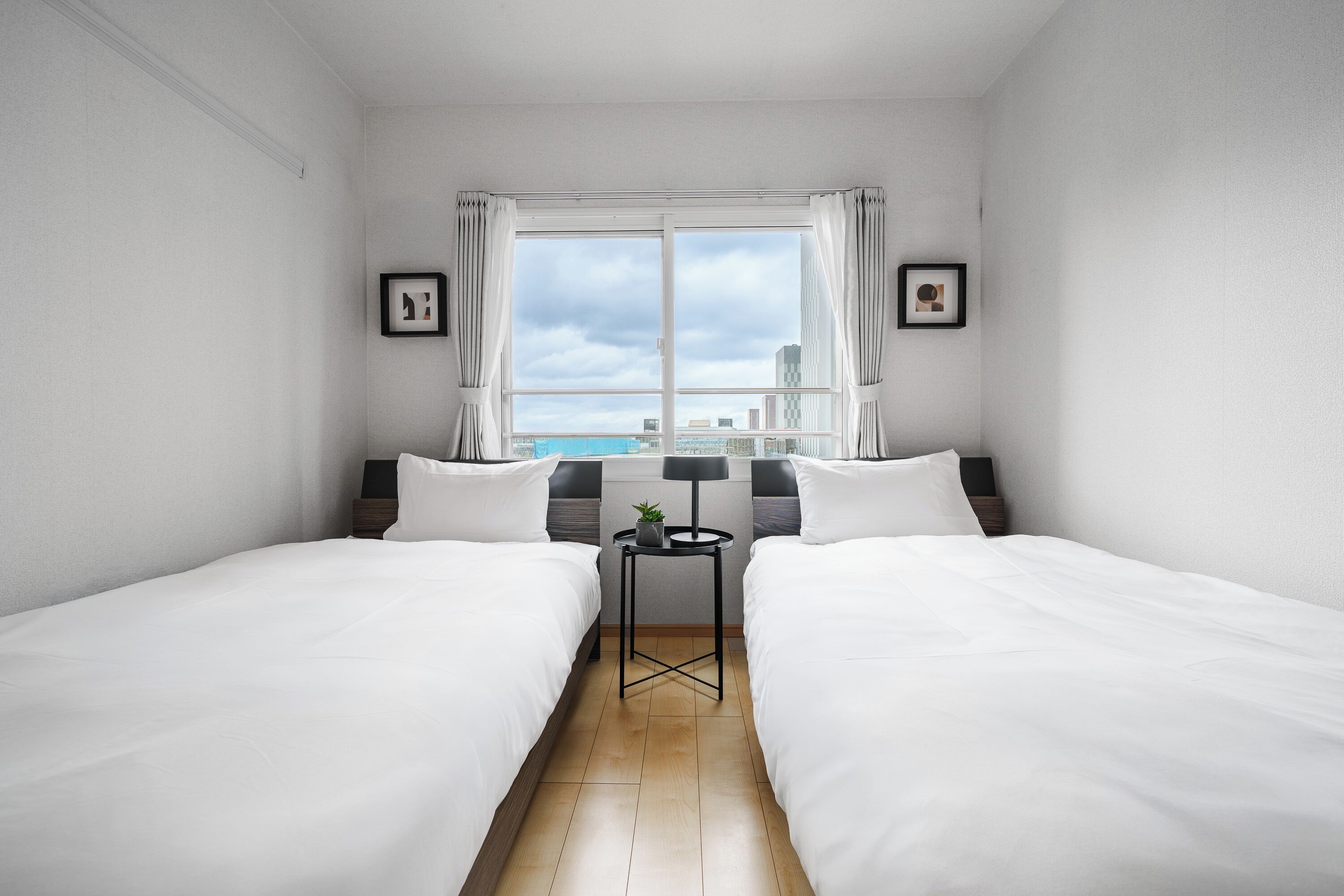 A hotel room with two beds, a small table, and a window with a city view.