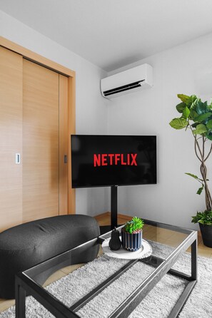 A modern living room with a flat-screen TV displaying 'NETFLIX' in red letters, a black sofa, a glass coffee table with a plant, and a wooden wardrobe. - E-US NAEBO STATION 408 (Sapporo)