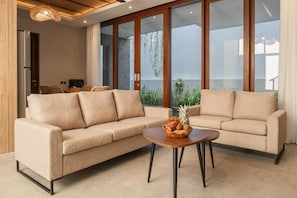 A modern living room with a beige sofa, a coffee table with fruit, and large windows with plants. - Solaris Canggu Villa by Tasvan (Badung)