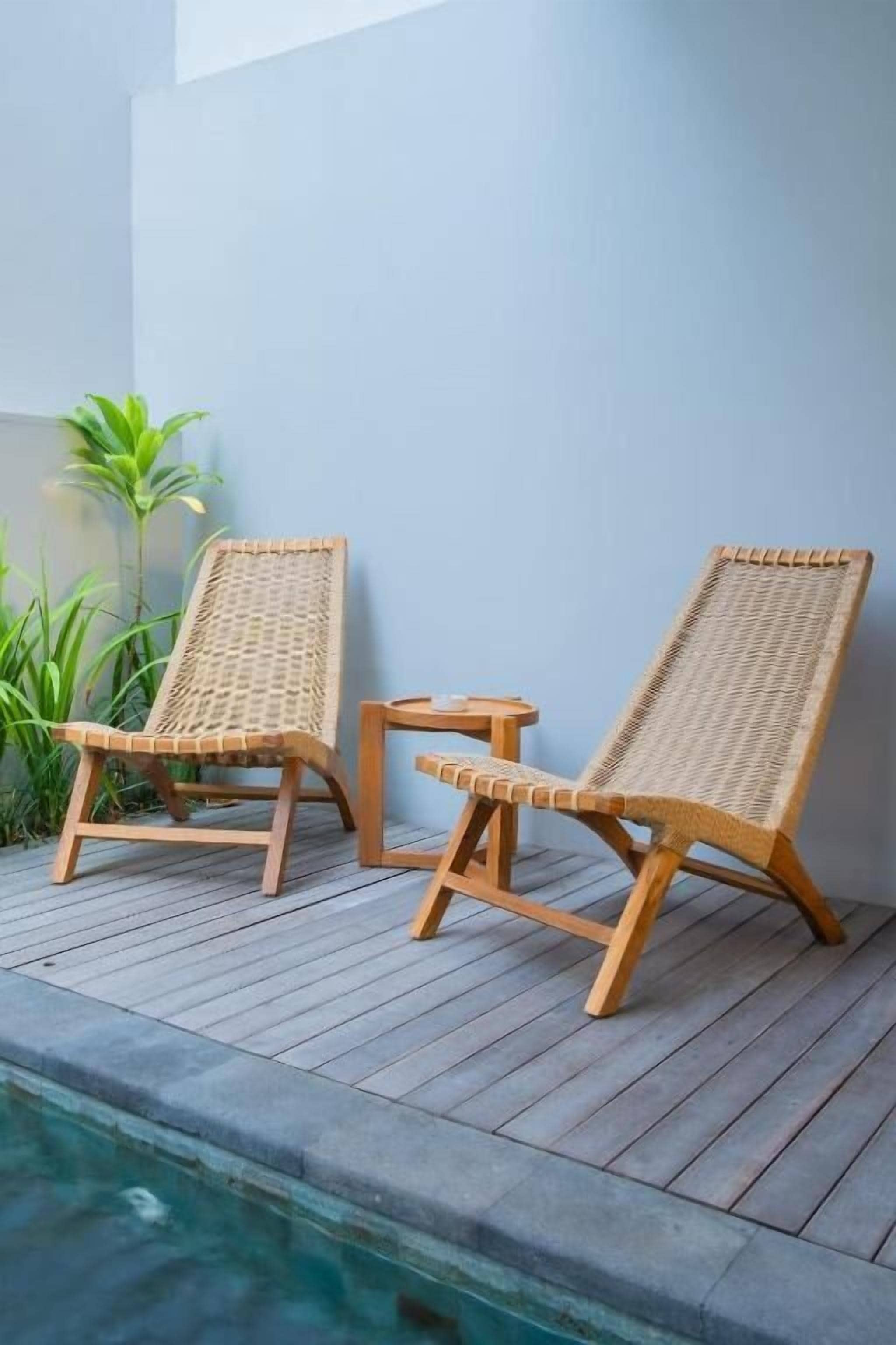 Two wicker lounge chairs with a small table on a wooden deck by a pool.