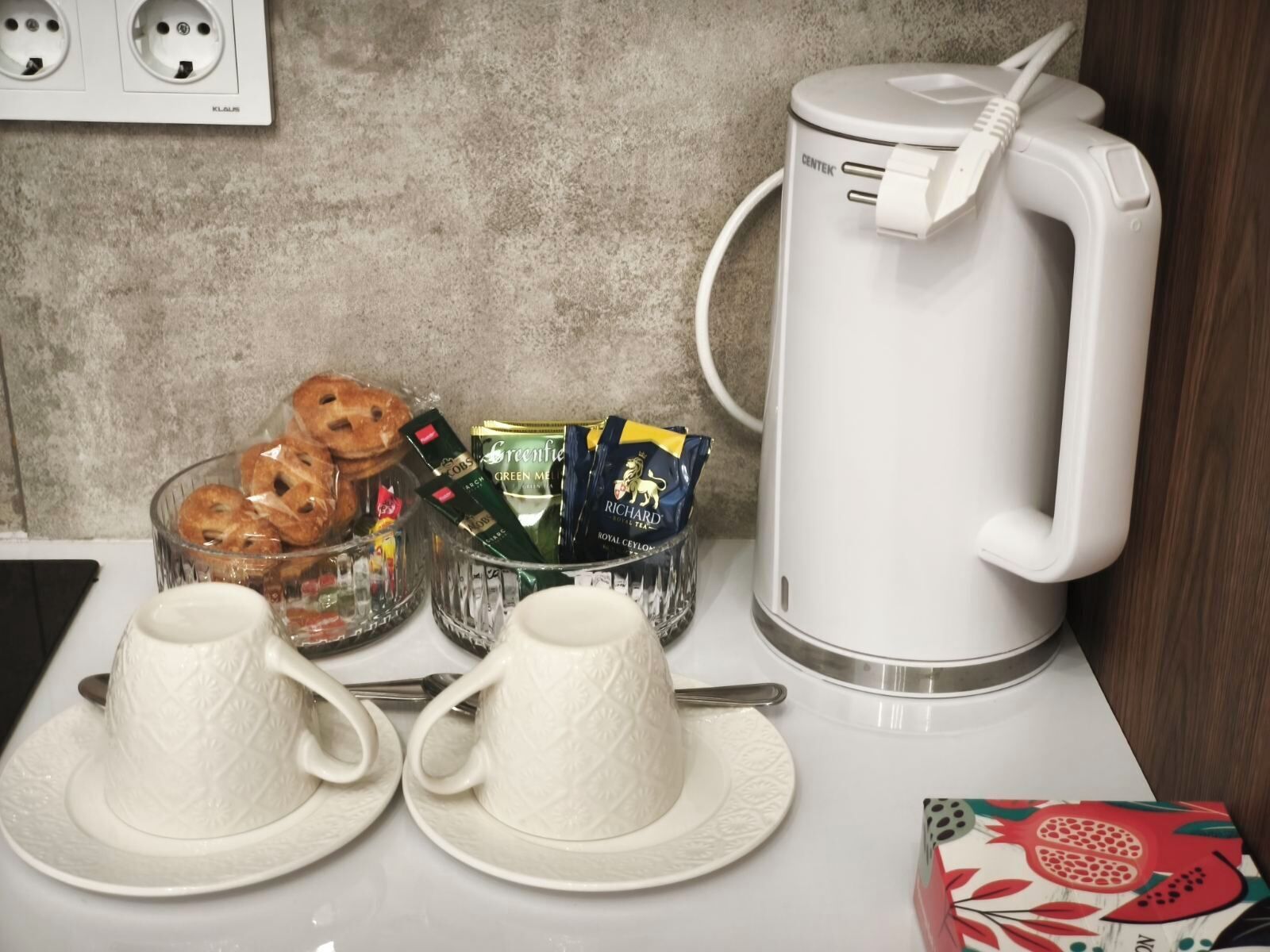 A kitchenette with a coffee maker, a bowl of pastries, and a box of tea.