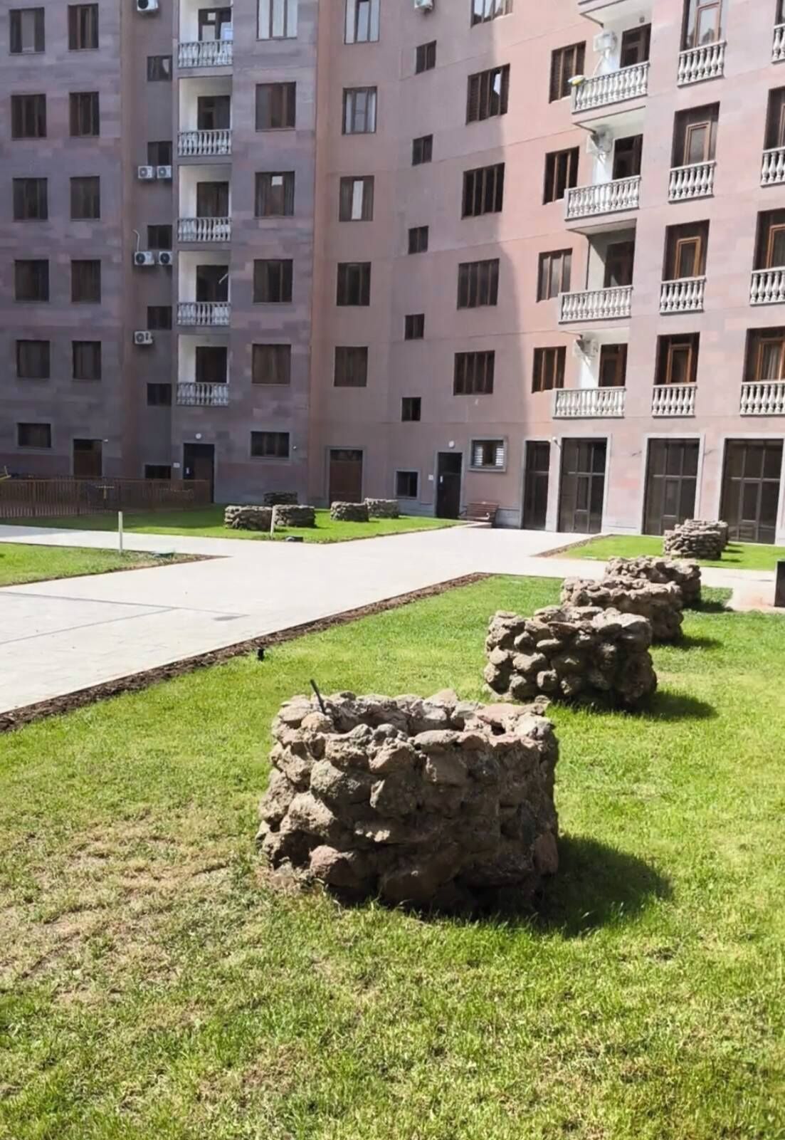 Edifício residencial com vários andares, varandas, um caminho pavimentado e estruturas de pedra em primeiro plano.