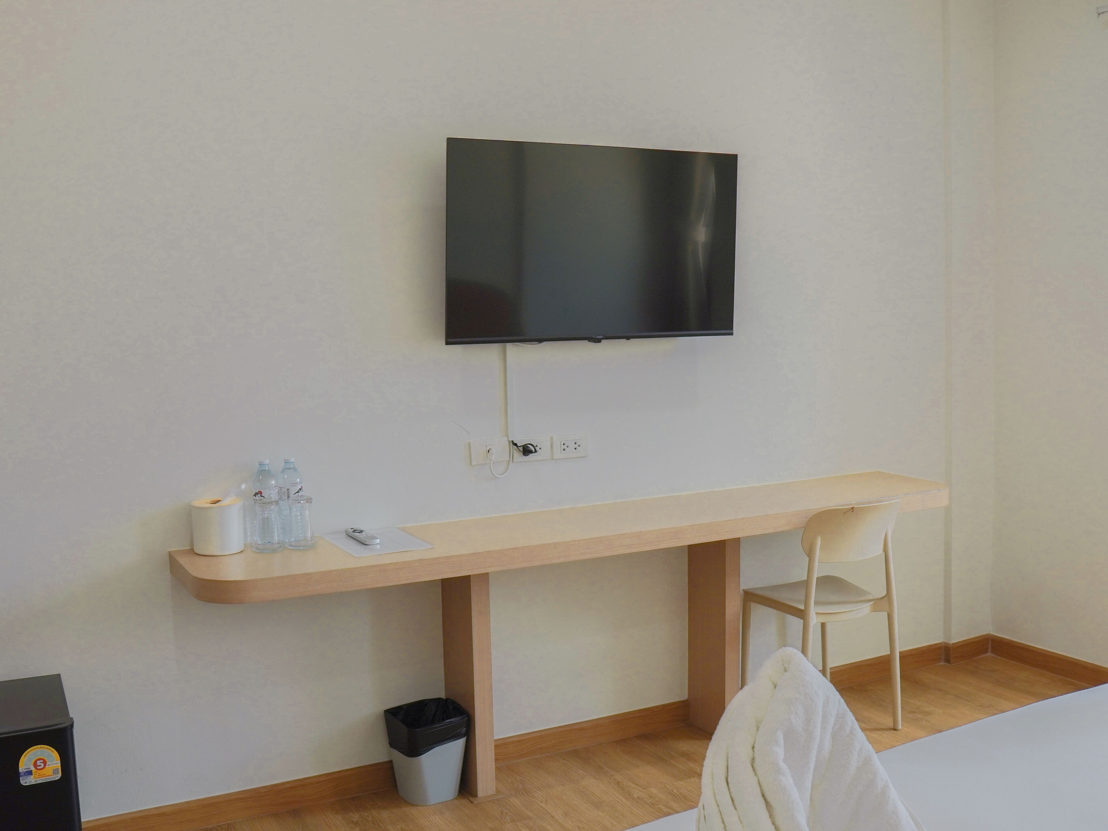Une pièce comprenant un bureau en bois, une chaise, un téléviseur fixé au mur, une poubelle et un petit objet noir posé sur le sol.