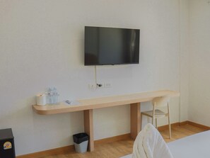 A room with a wooden desk, a chair, a television mounted on the wall, a trash bin, and a small black object on the floor.