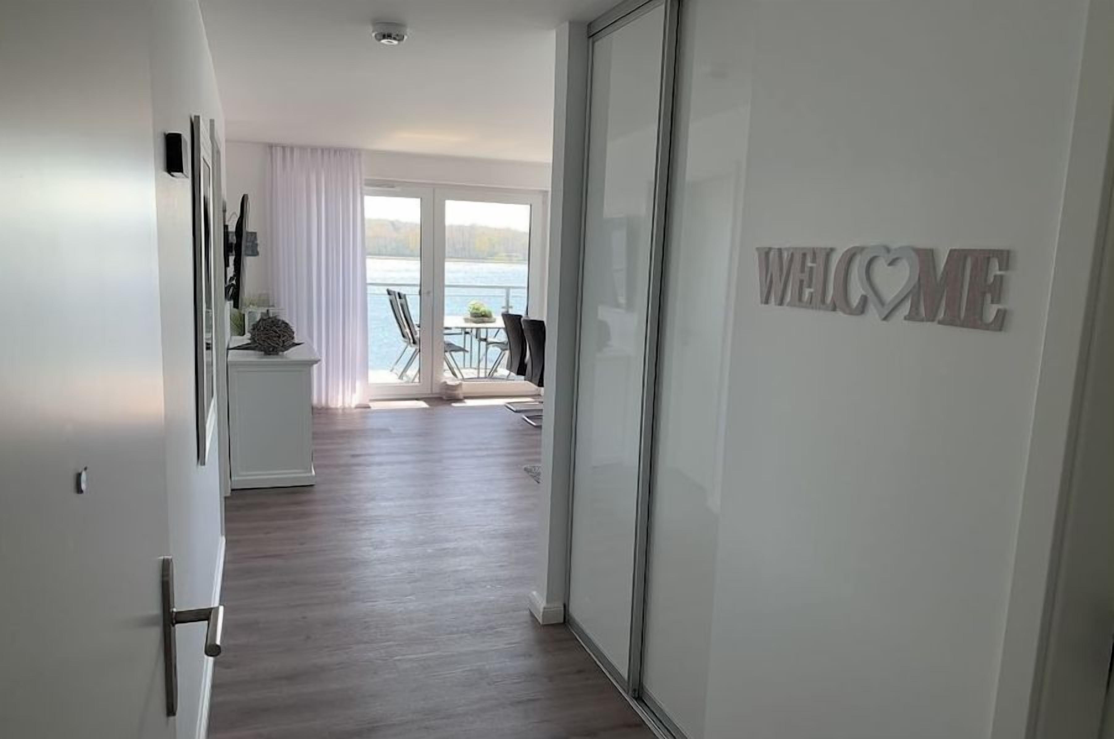 Un couloir menant à un balcon orné d’une pancarte « BIENVENUE », un coin repas avec des chaises et une table, et une vue sur une étendue d’eau.