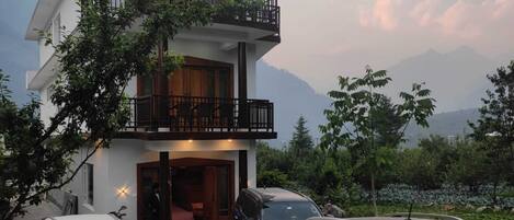 A modern two-story building with a balcony, parked cars, and a mountainous backdrop.