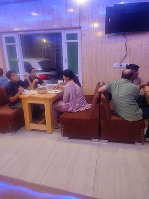 Un groupe de personnes est assis autour d’une table dans une pièce aux murs en bois, où un téléviseur est fixé au mur.