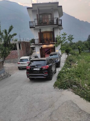 Un immeuble de deux étages doté d’un balcon, avec des voitures garées devant et une chaîne de montagnes en arrière-plan.
