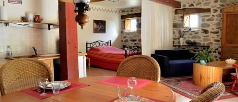A cozy room with a dining table set for two, a bed with a red bedspread, a blue sofa, and a stone wall.