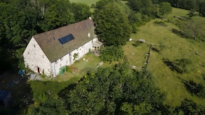Une maison en pierre avec un toit en pente et un panneau solaire, entourée d’une végétation luxuriante et avec des collines en arrière-plan.