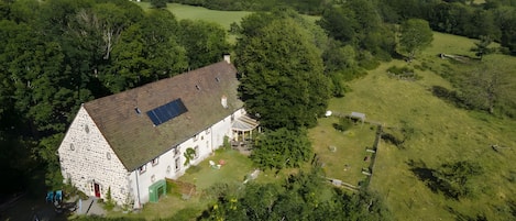 A stone house with a sloped roof and a solar panel, surrounded by lush greenery and hills in the background.