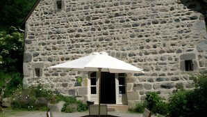 Un bâtiment en pierre avec un toit triangulaire, une petite fenêtre et une porte. Devant, on trouve une table ronde entourée de chaises et surmontée d’un parasol blanc.